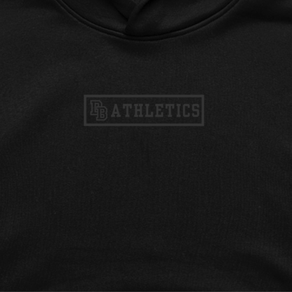 Close-up of The Issue Hoodie — black heavyweight fleece with matte black DB Athletics Brick vinyl emblem, minimal tone-on-tone design from the Varsity Drop Collection.